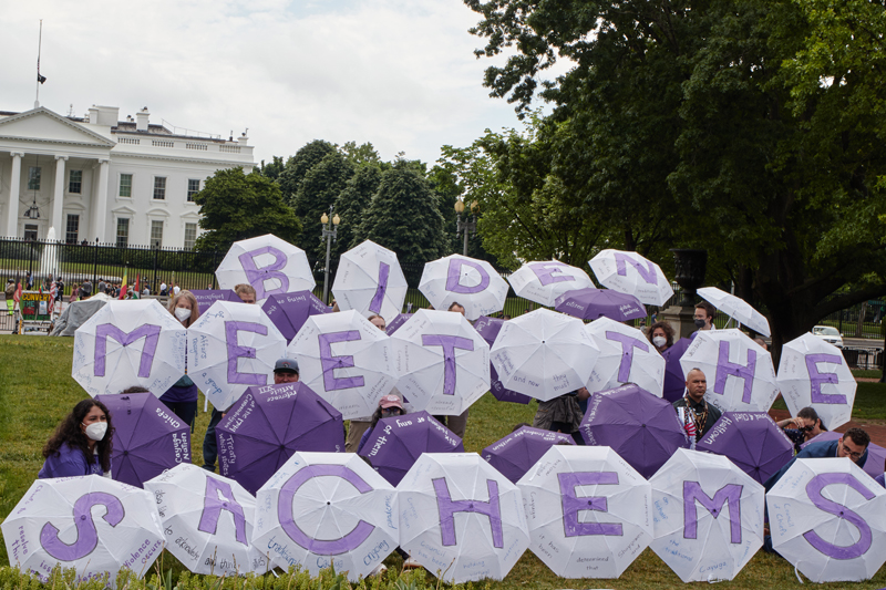 Photo of a group of people outside the White House, holding purple and white umbrellas with a message written on them that says Biden Meet The Sachems.