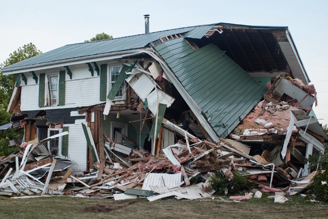 Photo of a house that has been half demolished.  Part of the roof has collapsed over a pile of broken beams and other material.