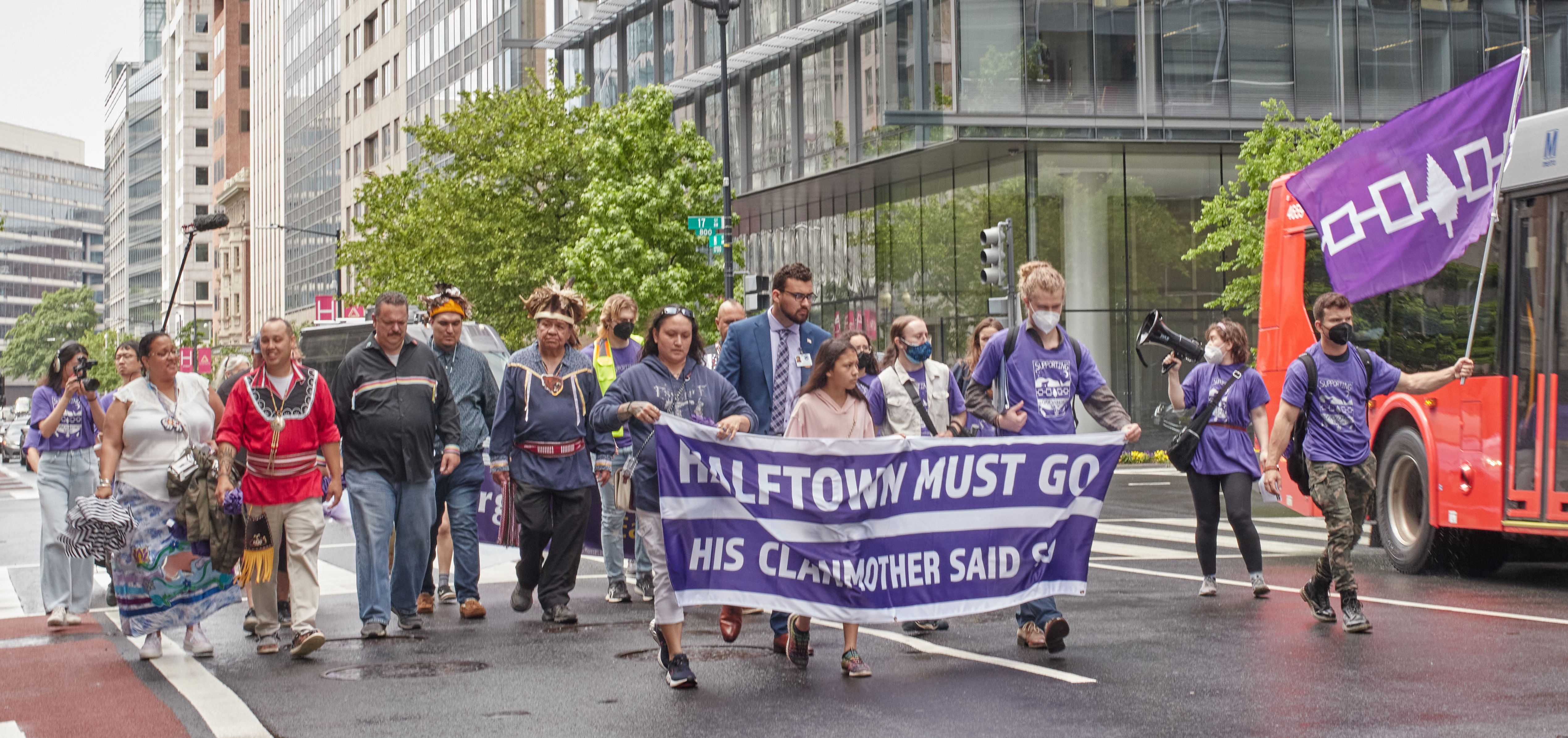 A photo of people marching in Washington, D.C. Some are holding a banner that says 'Halftown Must Go, His Clanmother Said So.' One person waves a large Haudenosaunee flag.